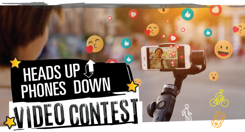 Graphic for the Heads Up Phones Down Video Contest, showing a pedestrian engrossed in the phone they are holding on a selfie stick. Emojis are coming out of the phone, blocking the pedestrian's view of where they are walking. The phone and emojis are in focus, while the real-world environment is blurry.